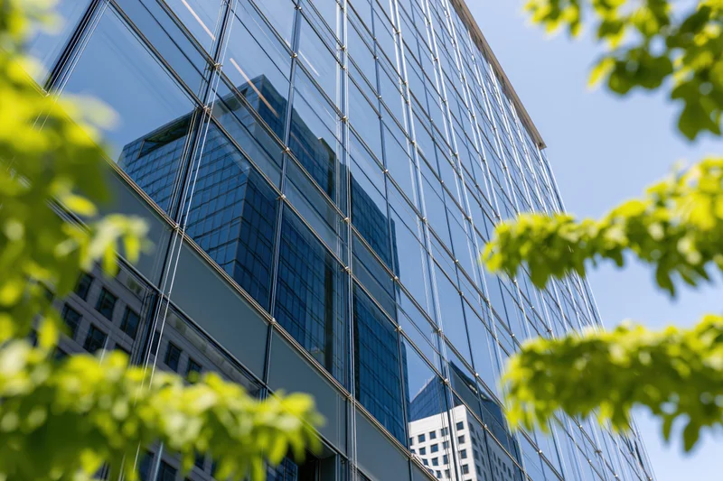Glass curtain wall of a modern office building reflecting sky and surrounding buildings, framed by green tree leaves in foreground