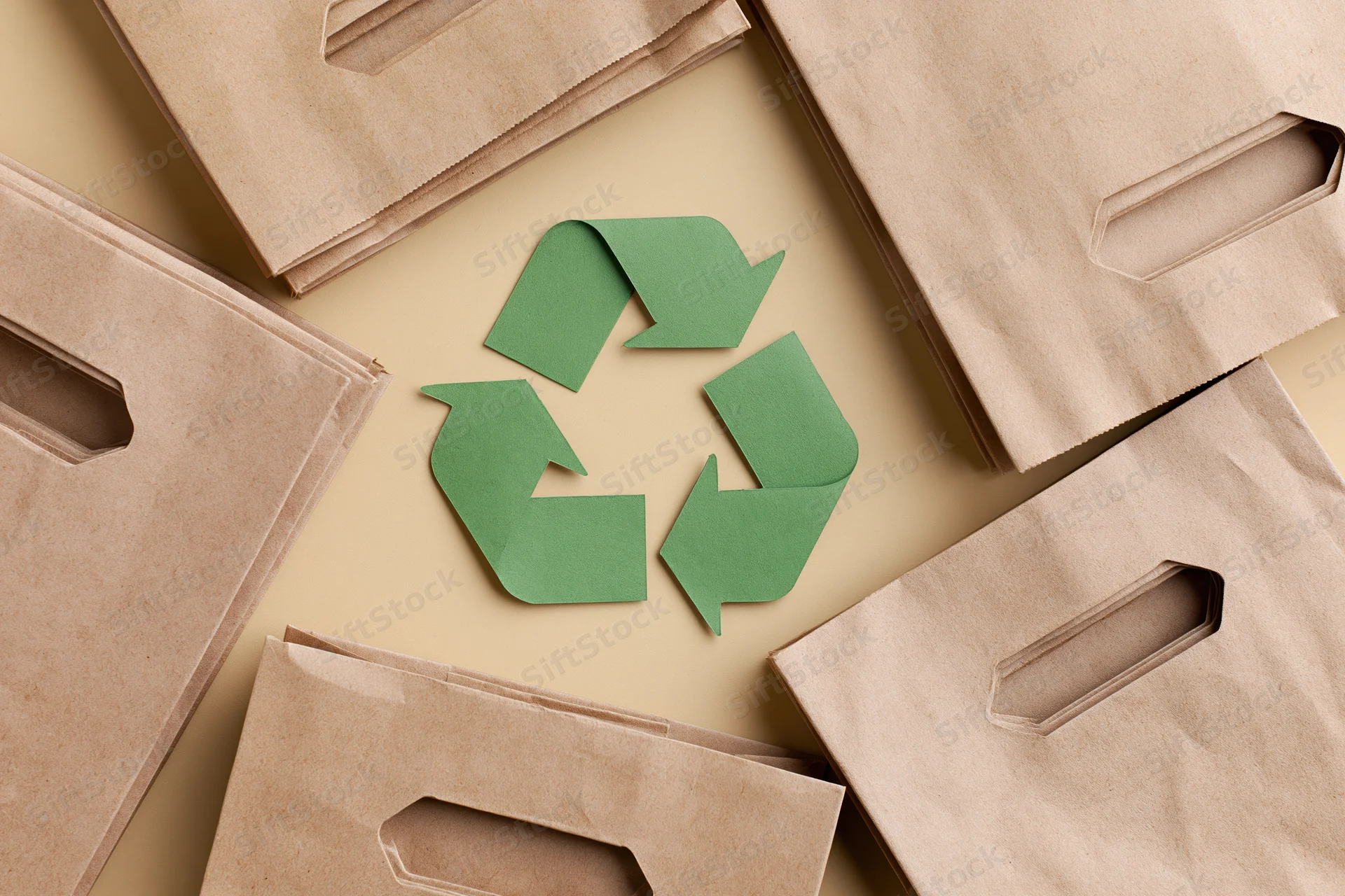 Green recycling symbol cut from paper lying flat at center surrounded by brown paper bags on beige surface