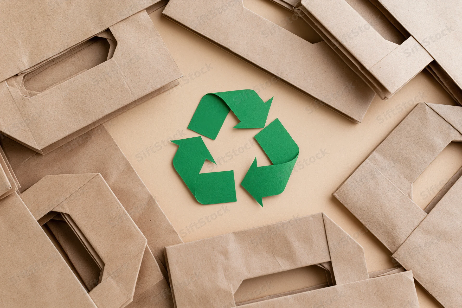 Green recycling symbol cut from paper lying flat at center surrounded by brown paper bags arranged in circular pattern on beige surface