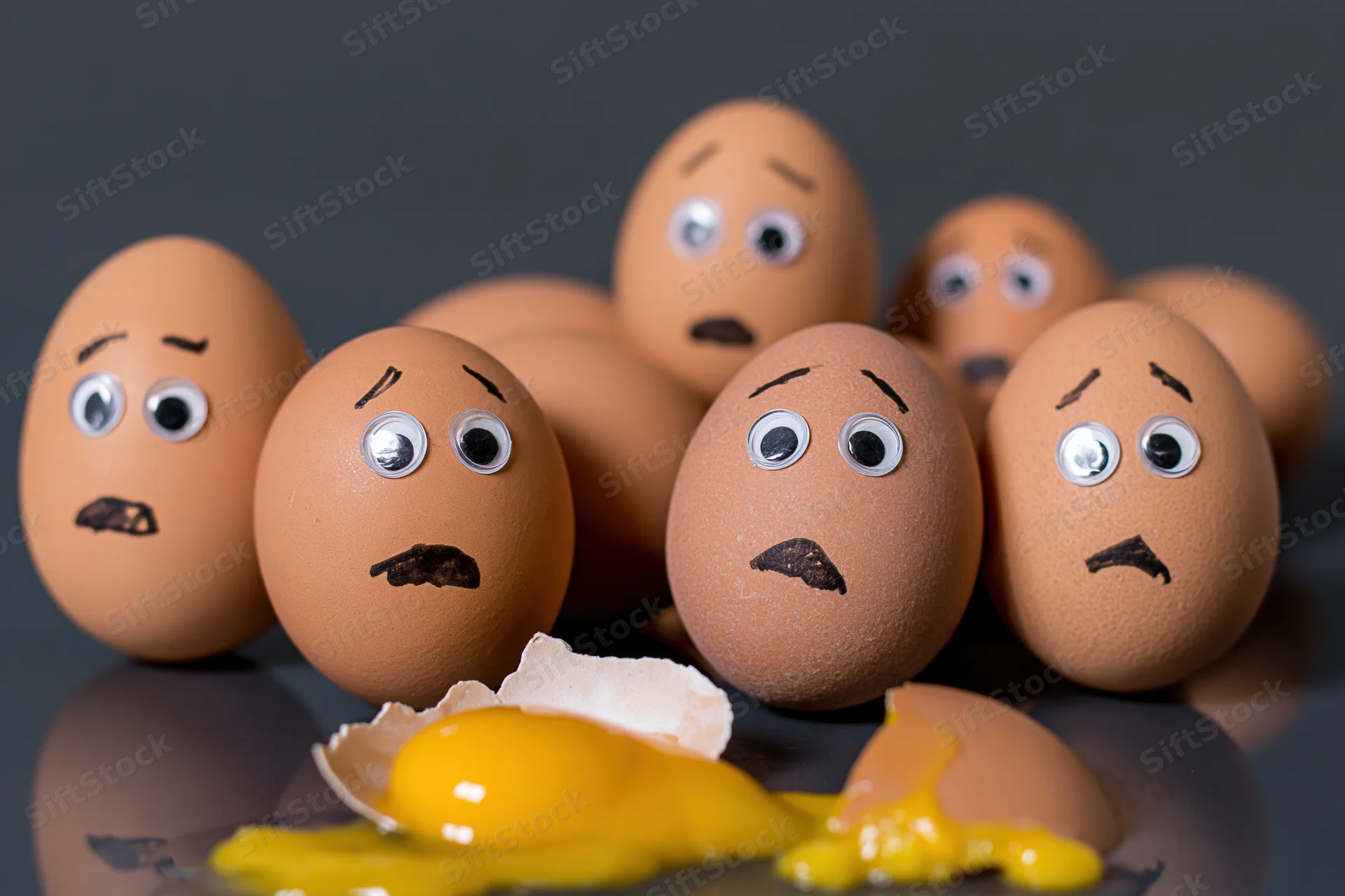 Group of brown eggs with googly eyes and marker-drawn faces gathered around a broken egg with yolk spilled on dark reflective surface