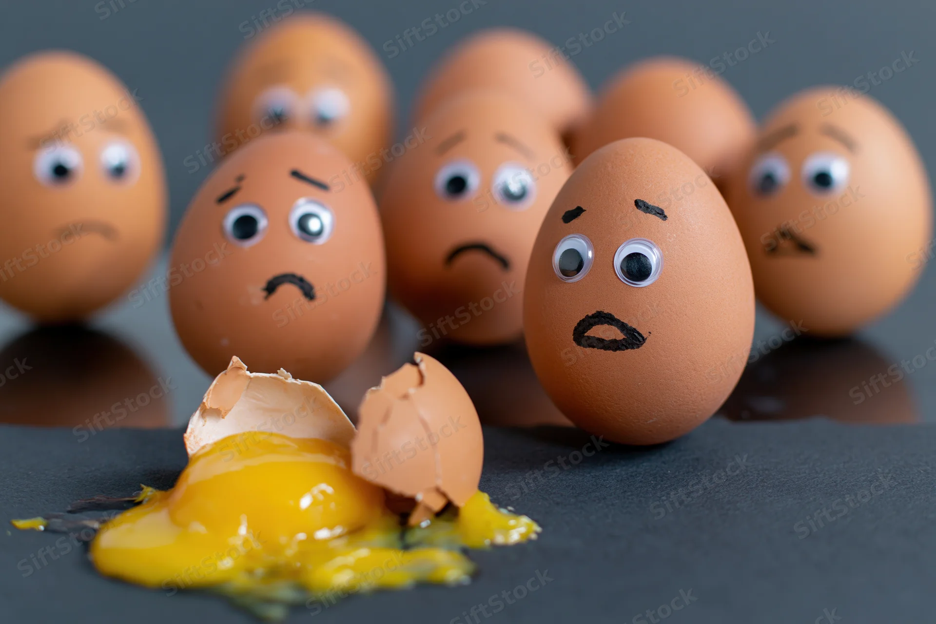 Group of brown eggs with googly eyes and marker-drawn faces reacting to a broken egg with spilled yolk on dark surface