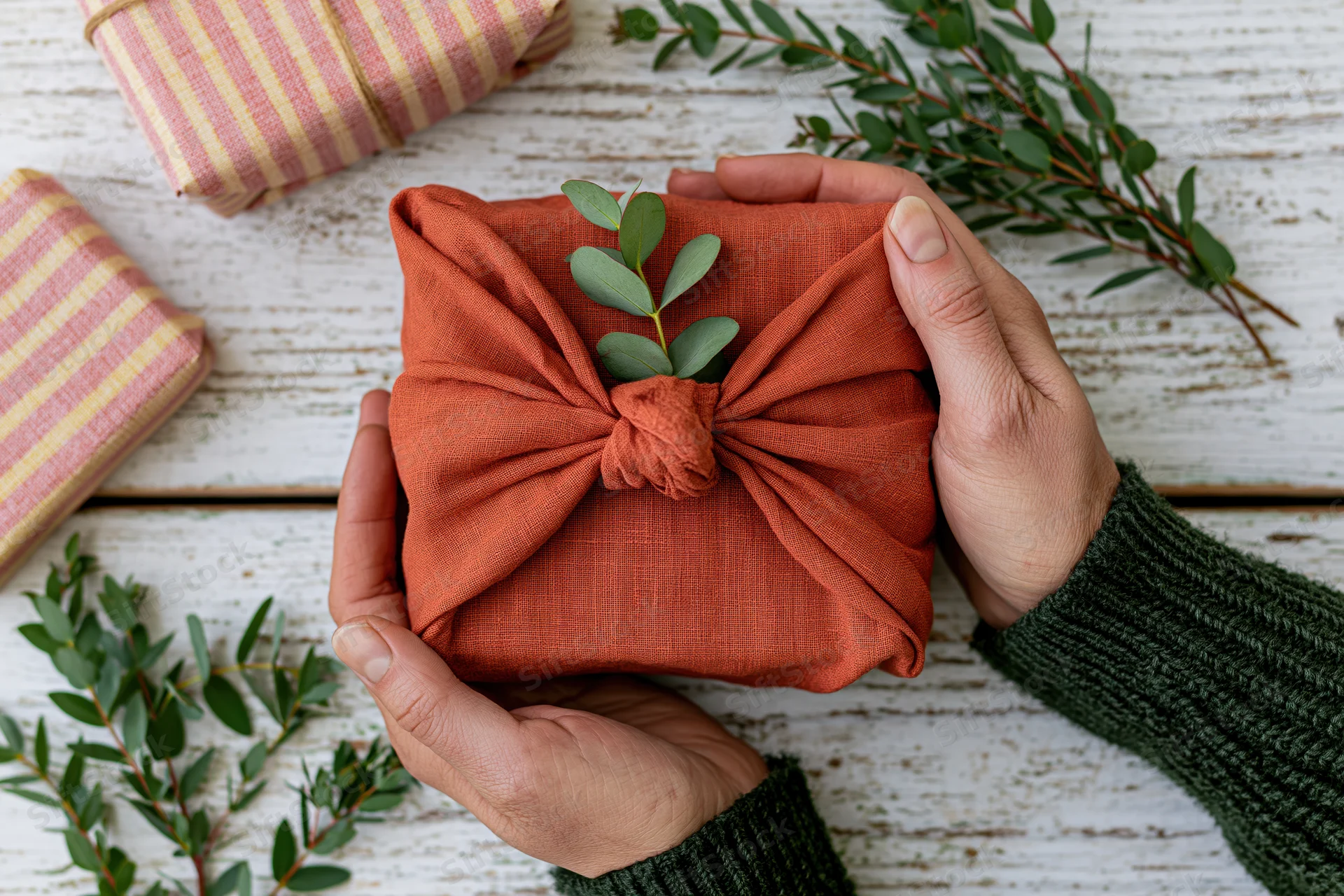 Hands holding a rust-orange fabric-wrapped gift with green eucalyptus sprig on white wooden surface