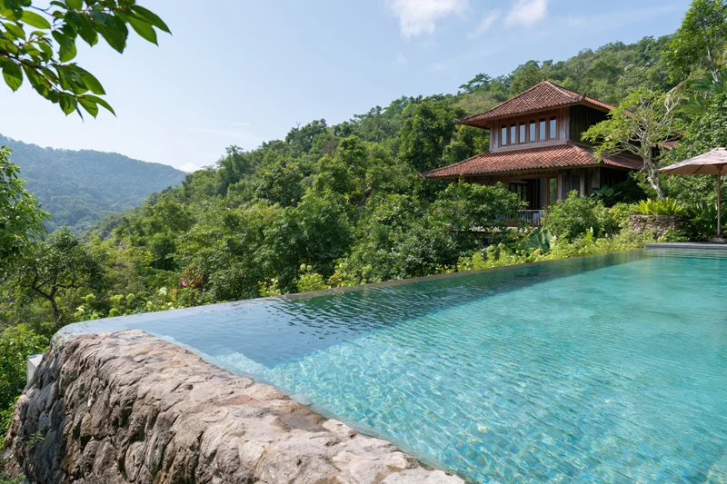 Infinity pool with natural stone edge and clear turquoise water overlooking lush tropical hills and a traditional villa
