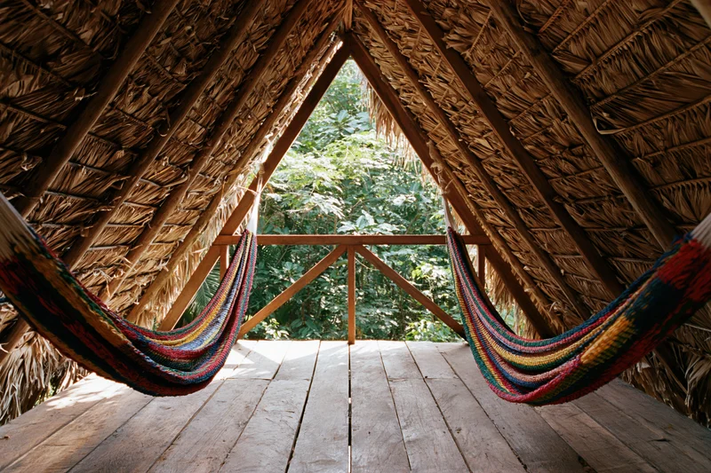 Interior of a thatched a-frame jungle hut with two colorful woven hammocks suspended from wooden beams, overlooking lush greenery