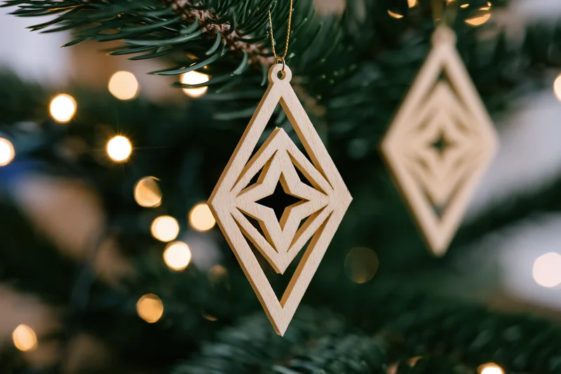 Laser-cut wooden diamond-shaped ornament hanging from a pine tree branch with warm bokeh lights in the background