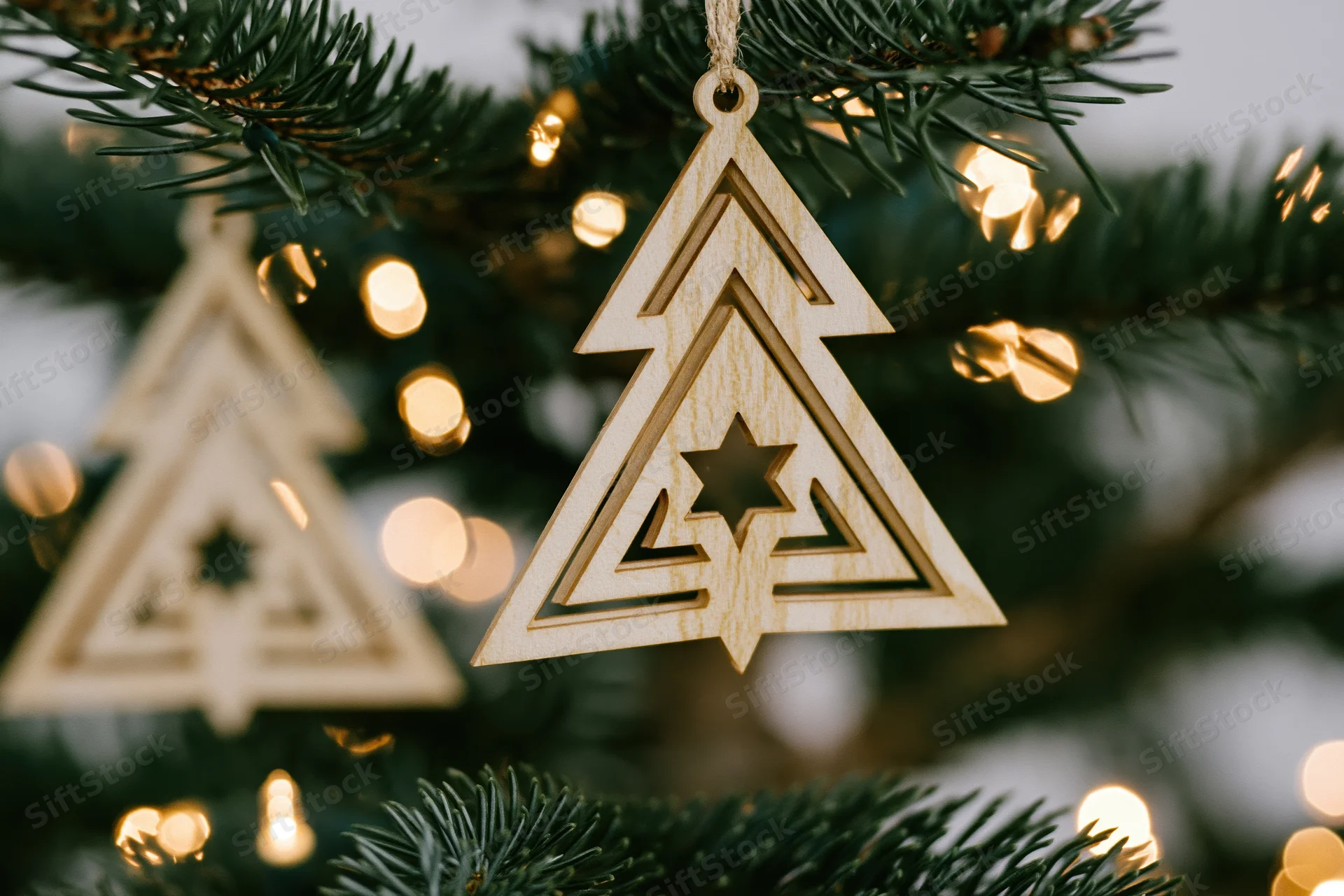 Laser-cut wooden christmas tree ornament hanging from a pine branch with warm bokeh lights in the background