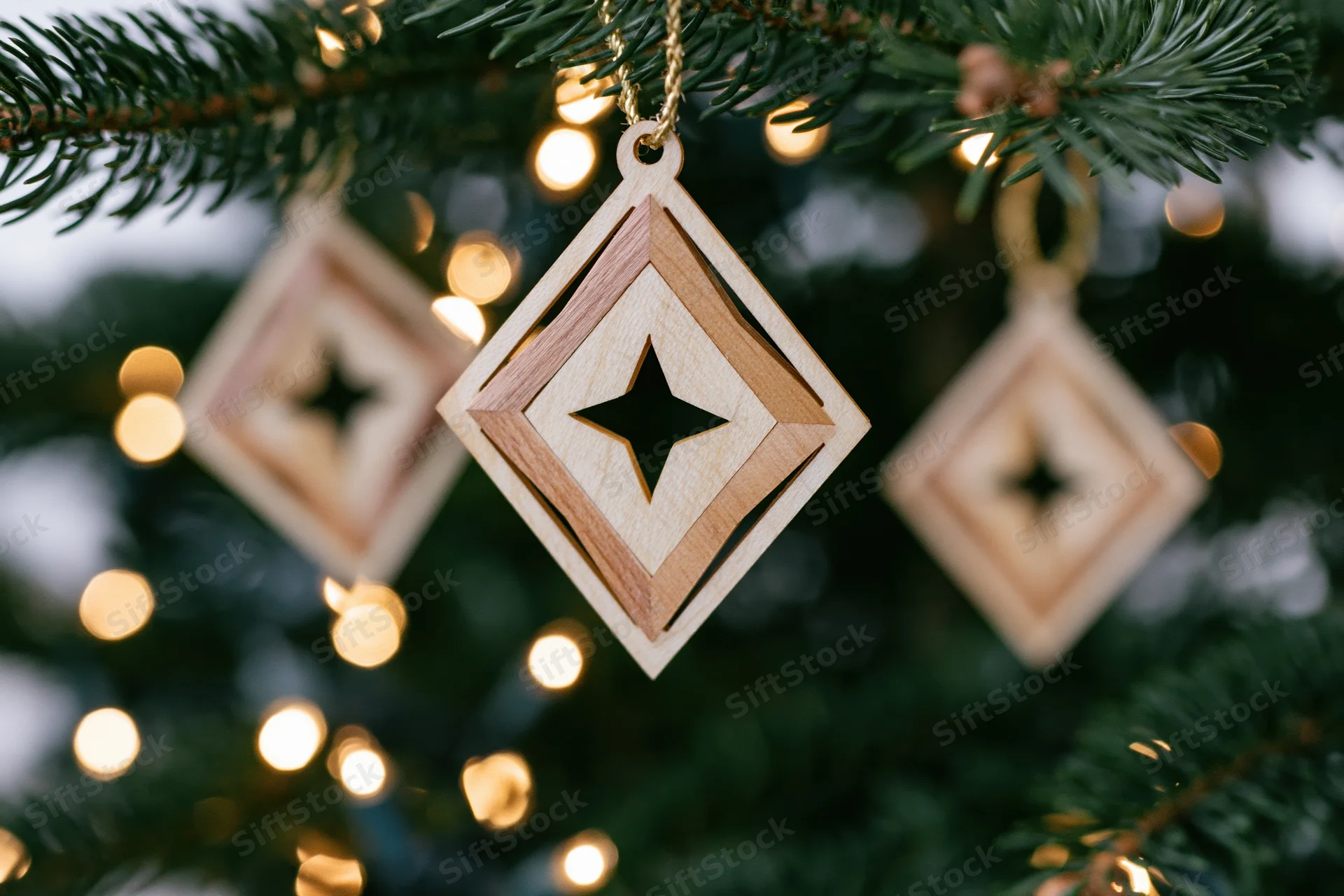 Laser-cut wooden diamond-shaped ornament with star cutout hanging from a pine tree branch with warm bokeh lights in the background