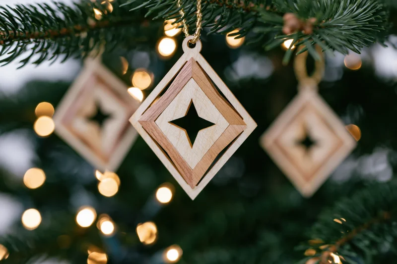 Laser-cut wooden diamond-shaped ornament with star cutout hanging from a pine tree branch with warm bokeh lights in the background