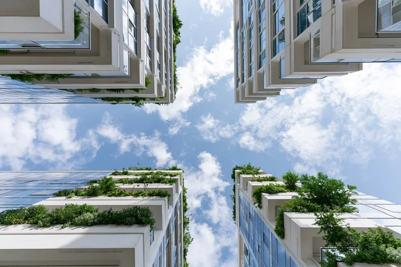 Modern glass-and-concrete skyscrapers with green rooftop gardens viewed from below against a blue sky with clouds