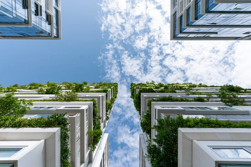 Modern skyscrapers with green vertical gardens viewed from below between buildings under blue sky