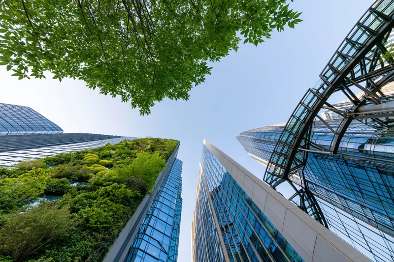 Modern glass skyscrapers with green vertical gardens viewed from ground level looking up between buildings under clear blue sky
