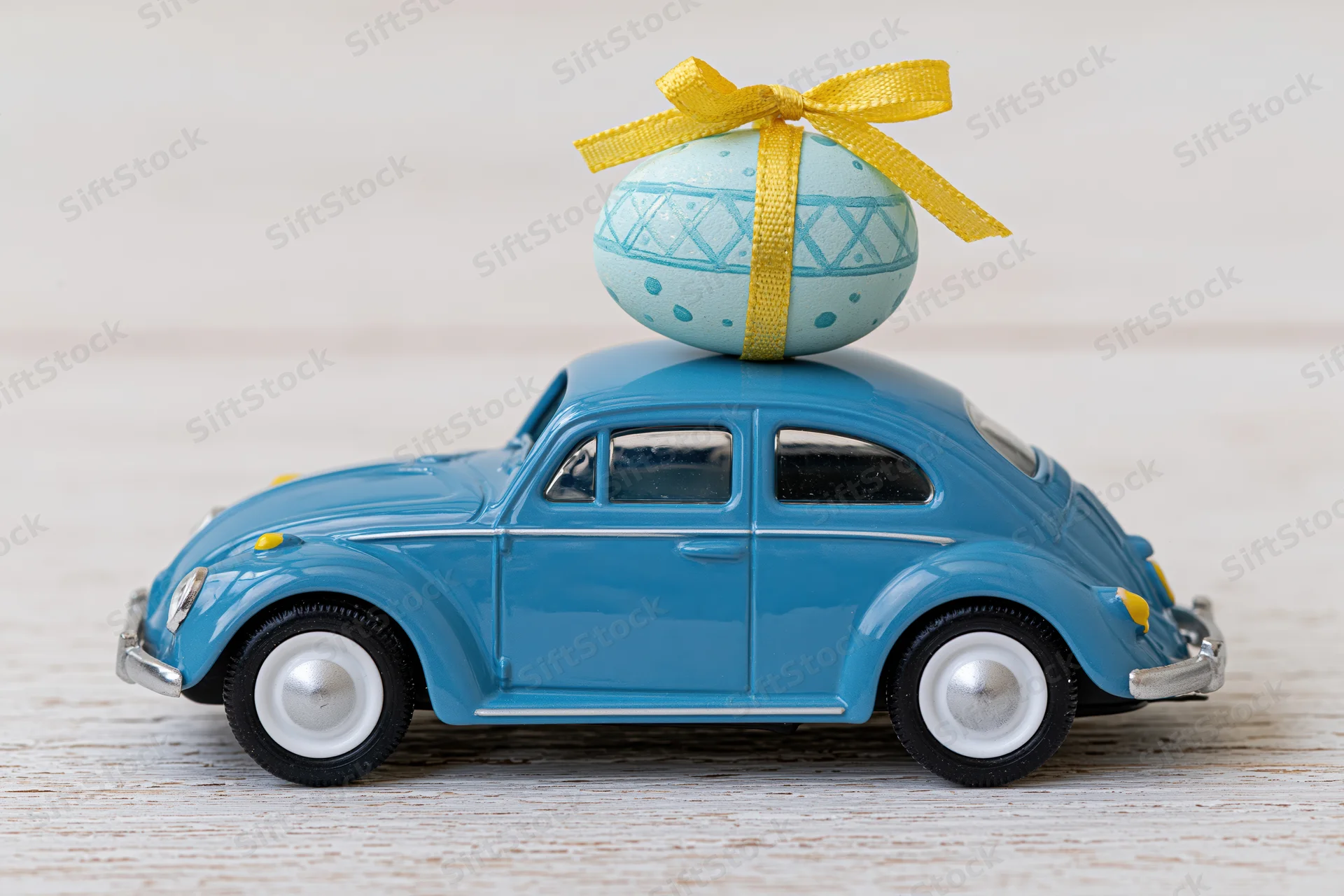Miniature blue toy volkswagen beetle carrying a decorated easter egg with yellow ribbon on its roof, placed on a light wooden surface