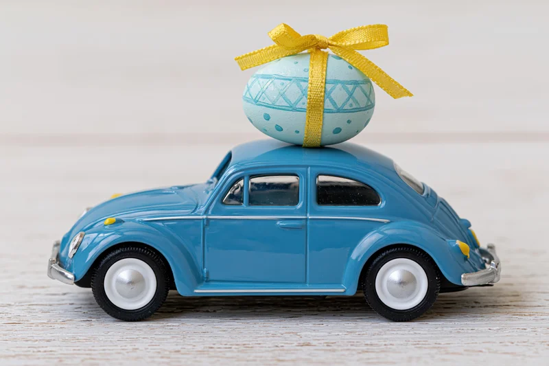 Miniature blue toy volkswagen beetle carrying a decorated easter egg with yellow ribbon on its roof, placed on a light wooden surface