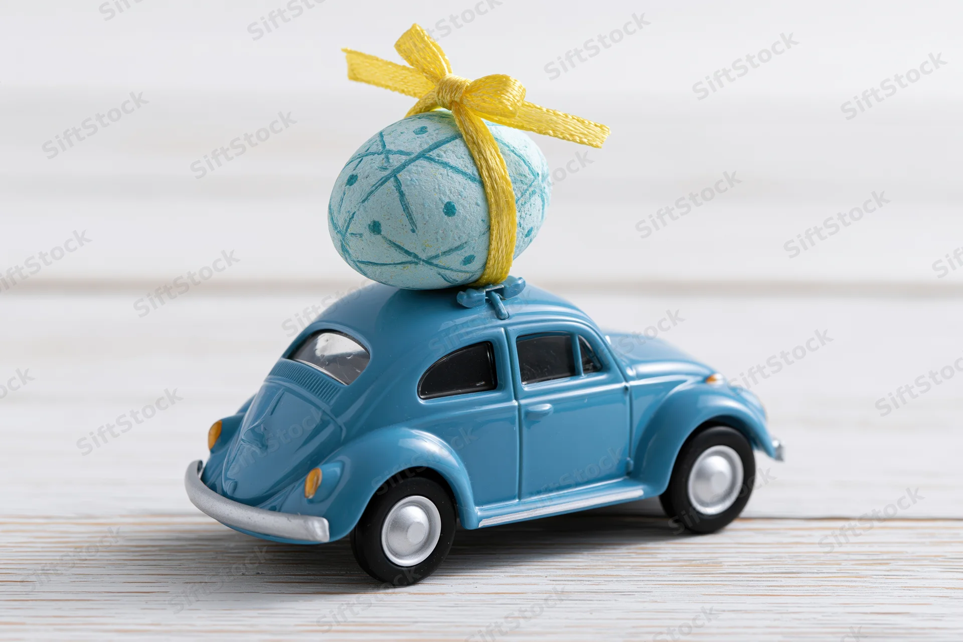 Miniature blue toy volkswagen beetle carrying a painted easter egg with yellow ribbon on its roof on a light wooden surface