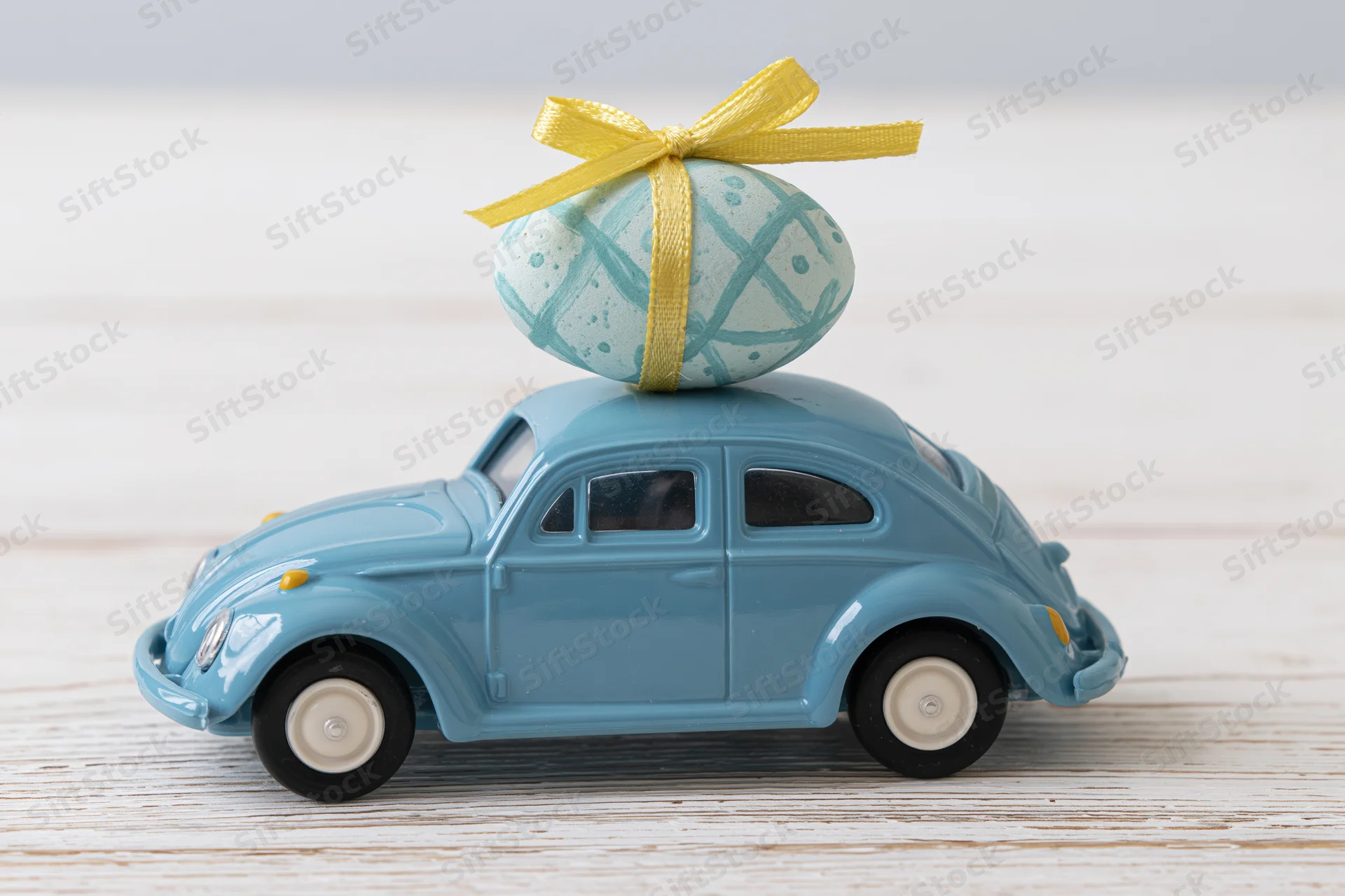 Miniature blue toy volkswagen beetle carrying a painted easter egg with yellow ribbon on its roof on a wooden surface