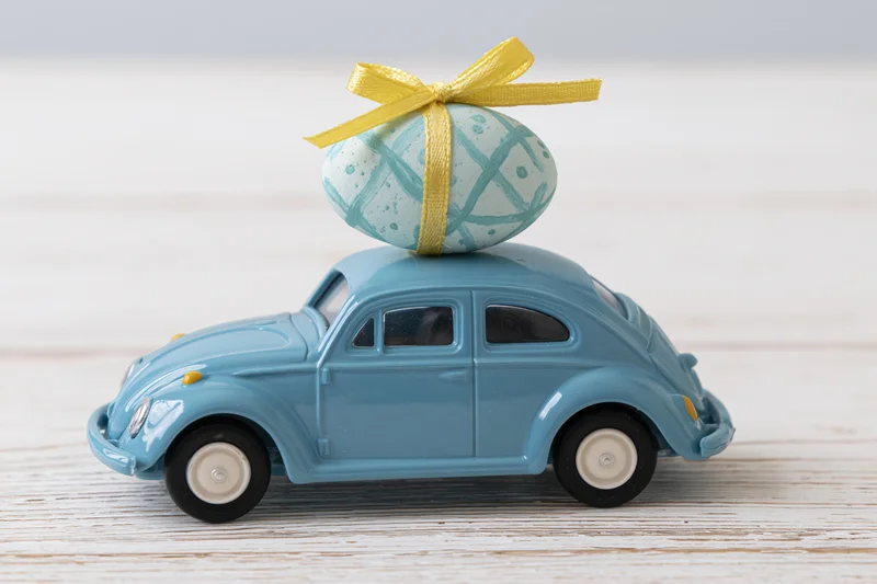 Miniature blue toy volkswagen beetle carrying a painted easter egg with yellow ribbon on its roof on a wooden surface