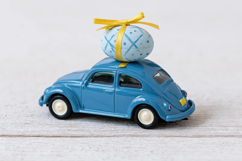 Miniature blue toy volkswagen beetle carrying a painted easter egg with yellow ribbon on a white wooden surface