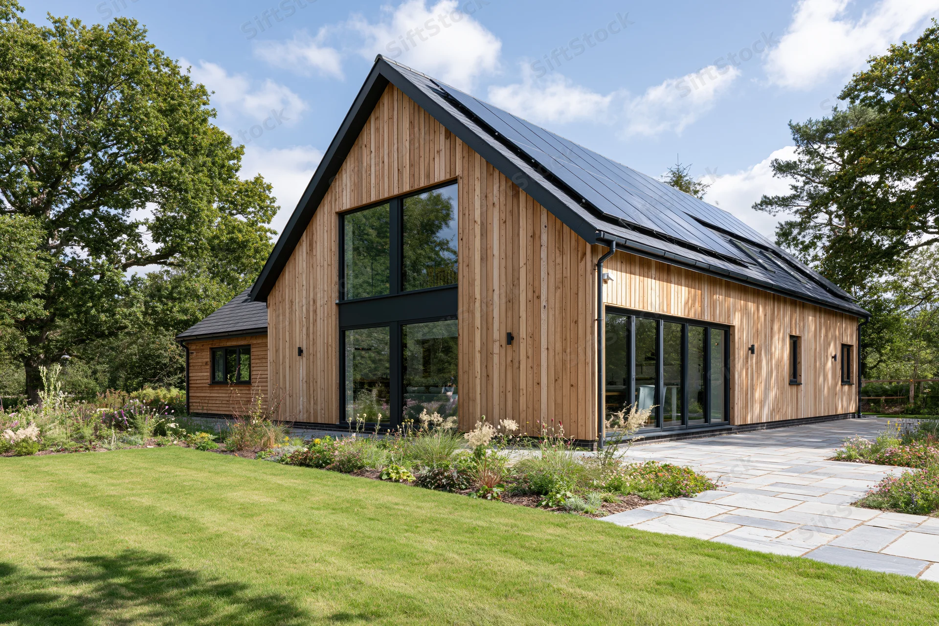 Modern barn-style house with vertical timber cladding and solar panels on the roof, surrounded by green lawn and trees under a blue sky with clouds