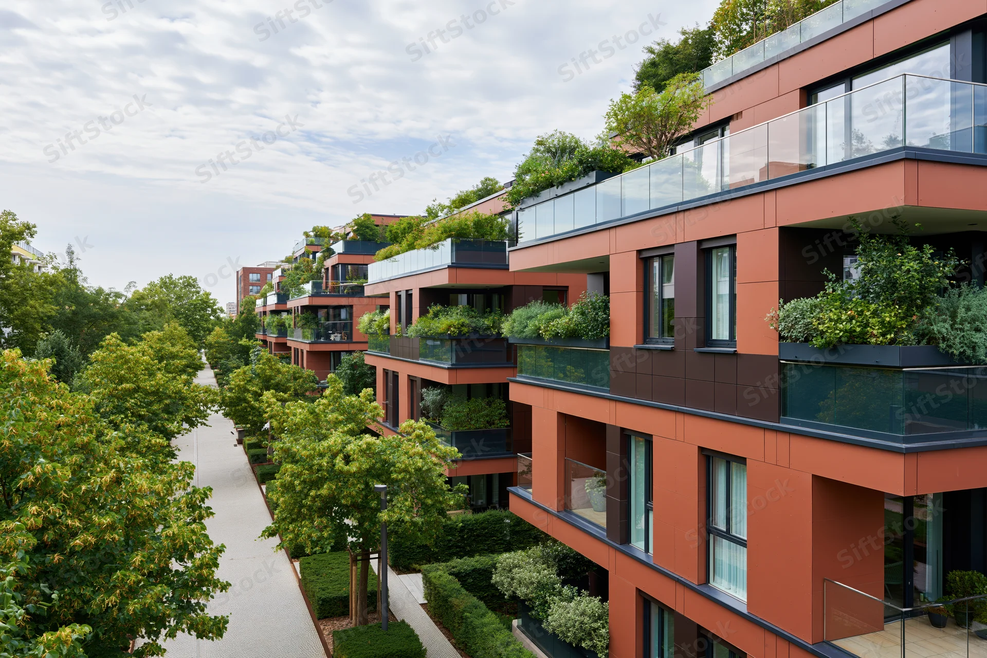 Modern residential apartment complex with rooftop gardens and balconies lined with greenery along a tree-lined pathway