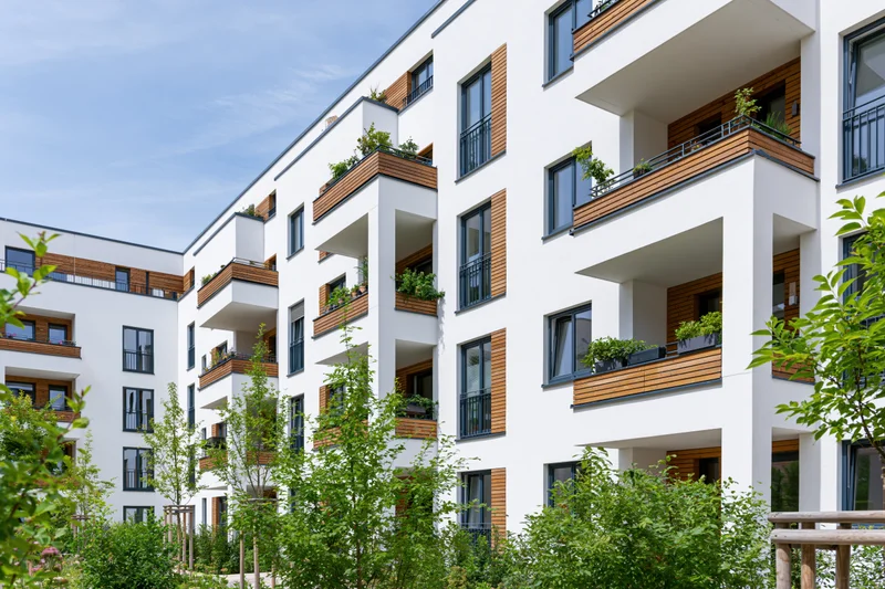 Modern white residential apartment building with wooden balconies and greenery in an urban courtyard setting