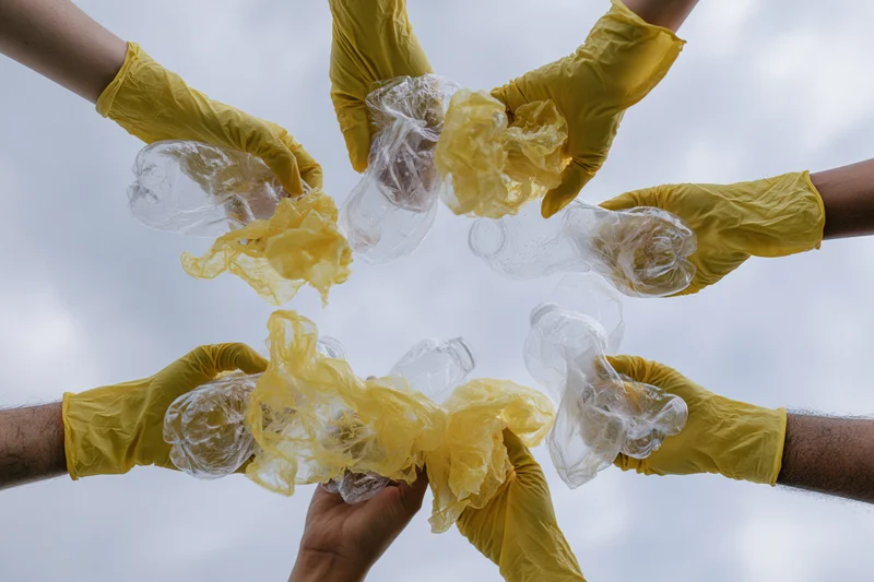 Multiple hands in yellow rubber gloves holding crumpled plastic bottles in a circle against a cloudy sky