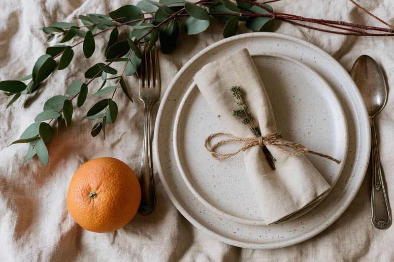 Rustic place setting arranged overhead on crumpled linen with ceramic plates, linen napkin tied with twine and thyme, silverware, orange, and eucalyptus branches