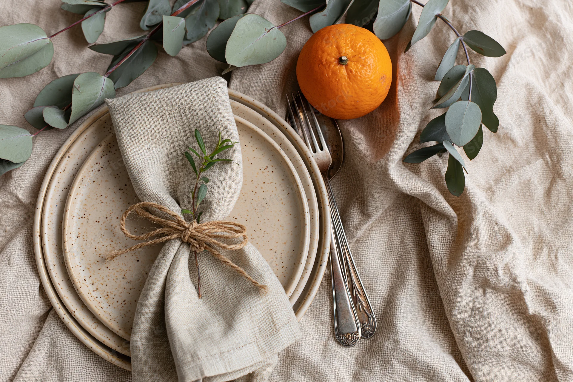 Rustic place setting with linen napkin, ceramic plates, cutlery, orange, and eucalyptus on crumpled linen tablecloth
