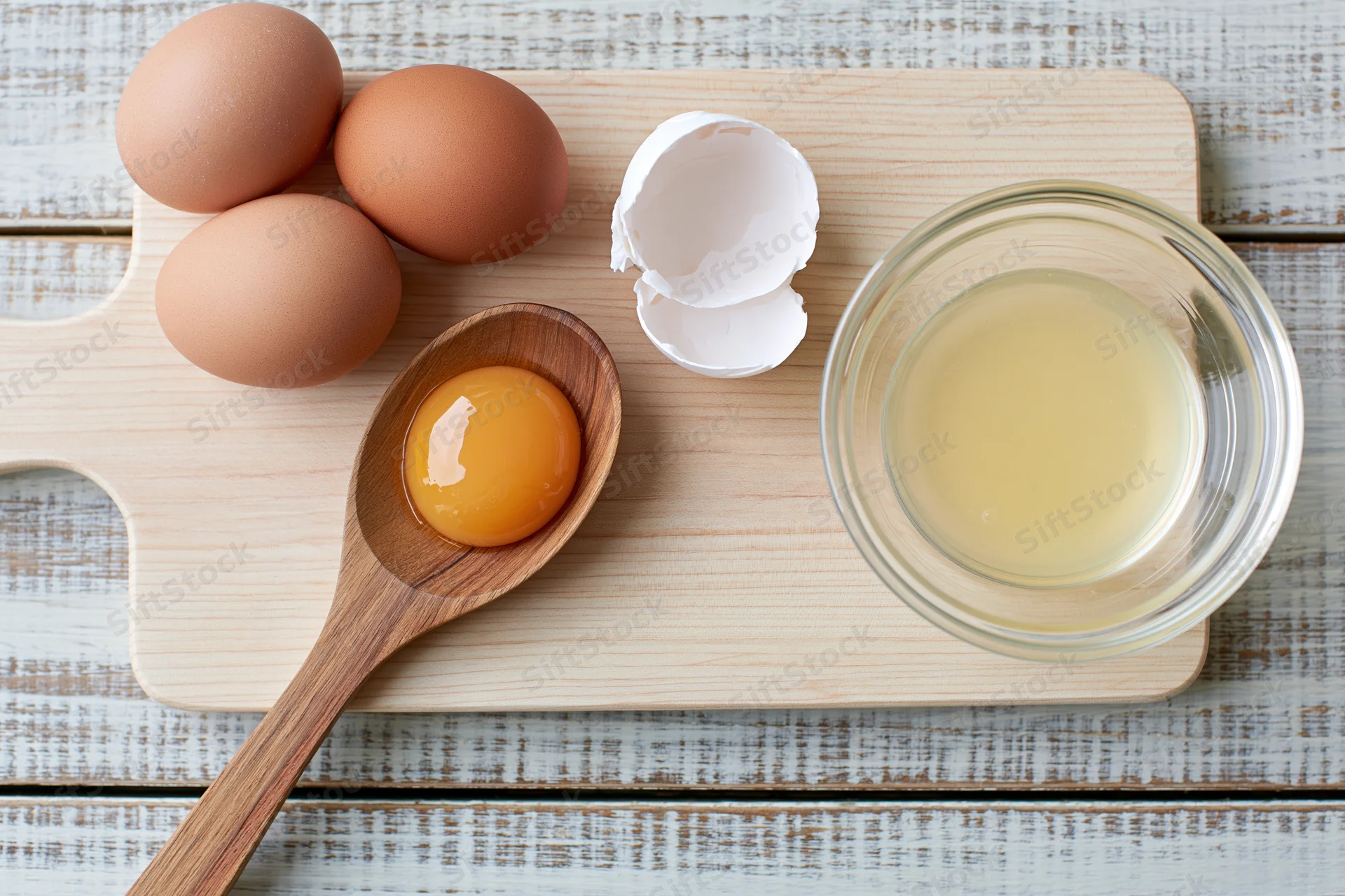 Raw egg yolk in wooden spoon on maple cutting board with whole eggs, shell, and bowl of egg white nearby