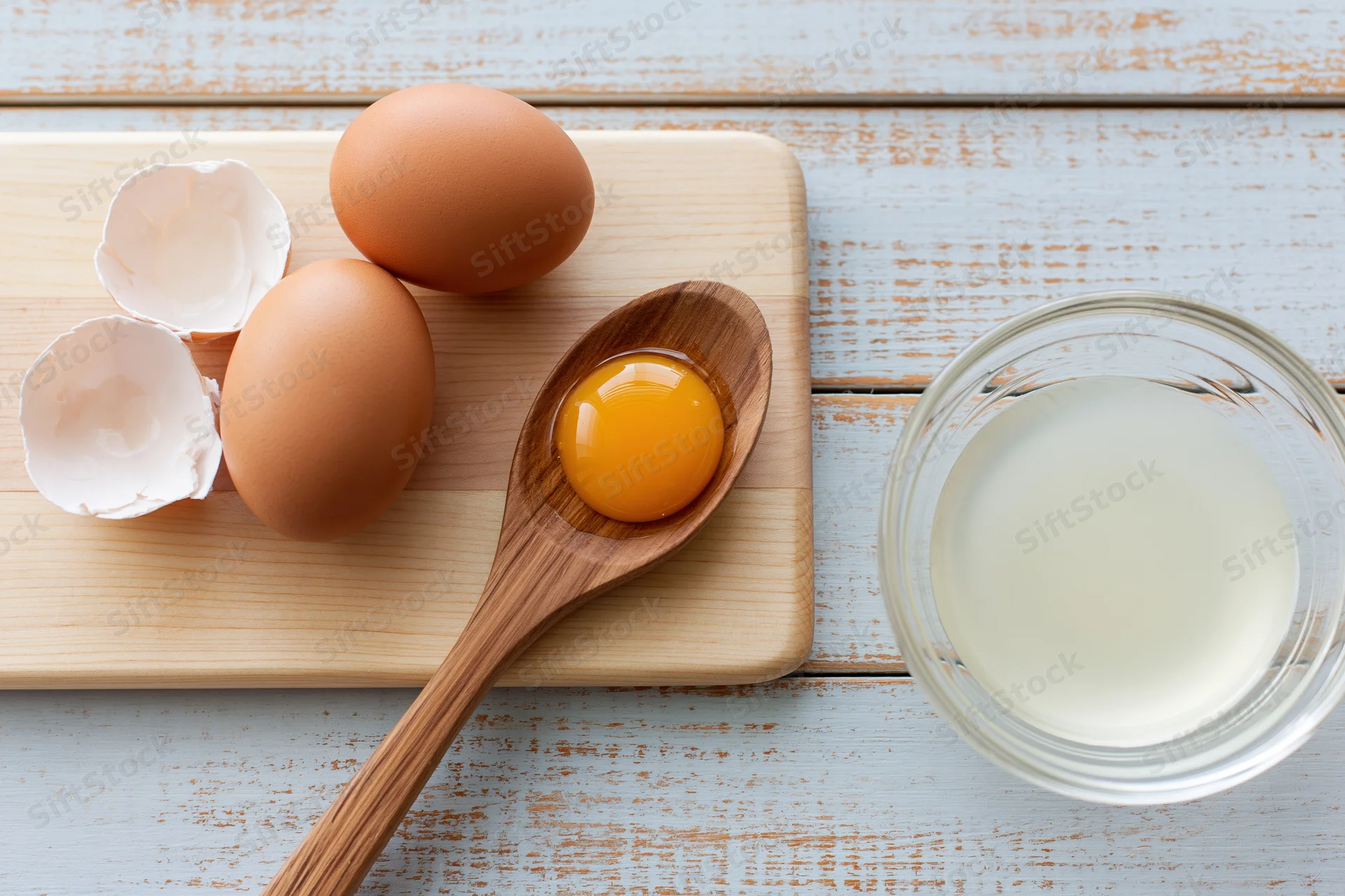 Raw egg yolk in wooden spoon on cutting board with whole eggs and milk nearby