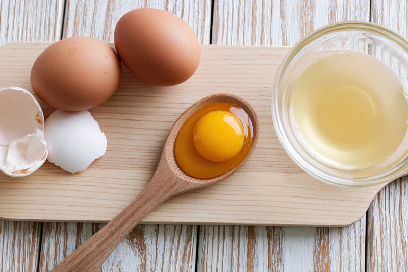 Raw egg yolk in wooden spoon on maple cutting board with whole eggs and eggshells nearby
