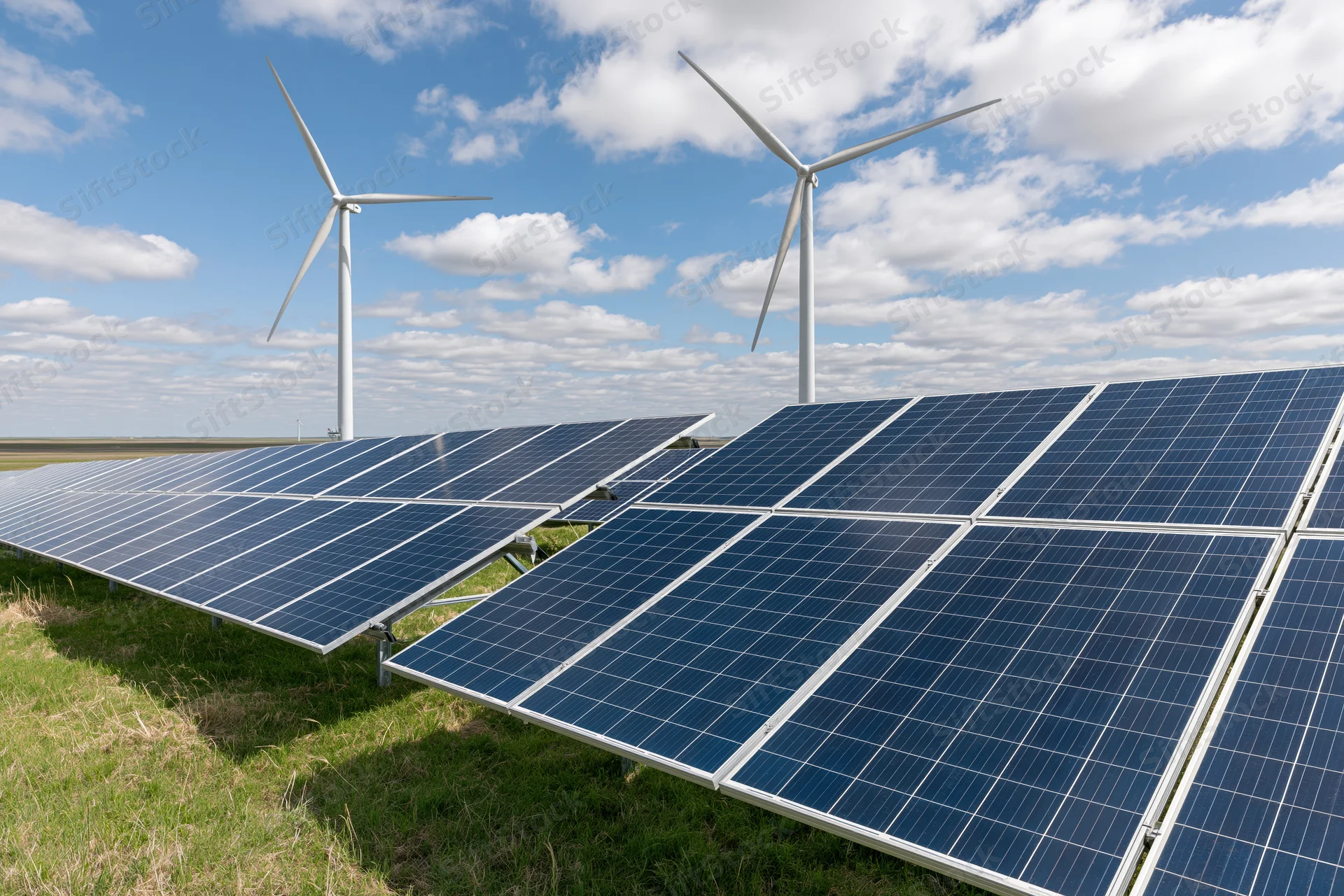 Rows of solar panels and wind turbines in a grassy field under a partly cloudy sky
