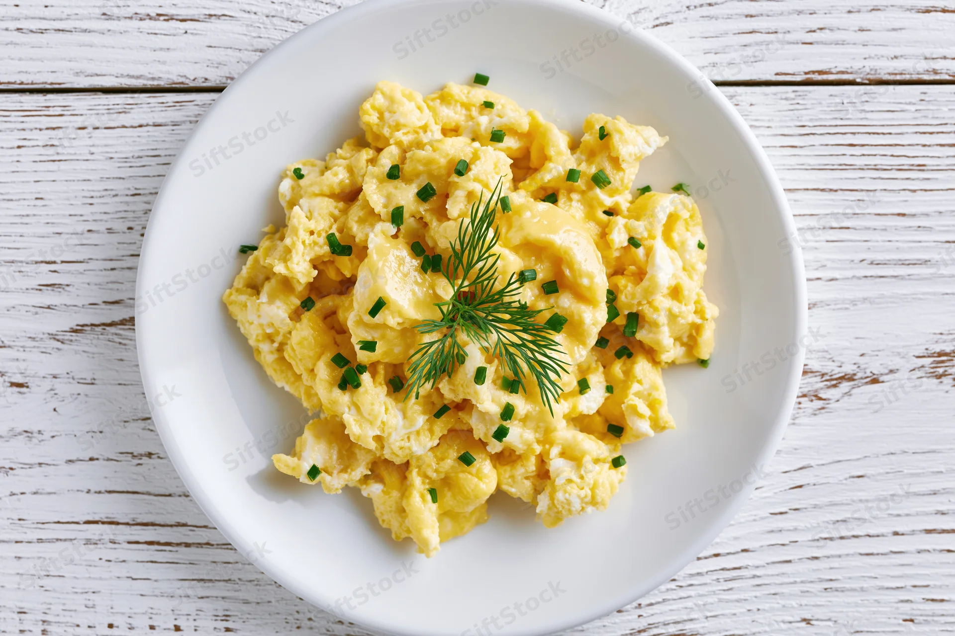 Scrambled eggs garnished with dill and chives on a white ceramic plate set on a whitewashed wooden table