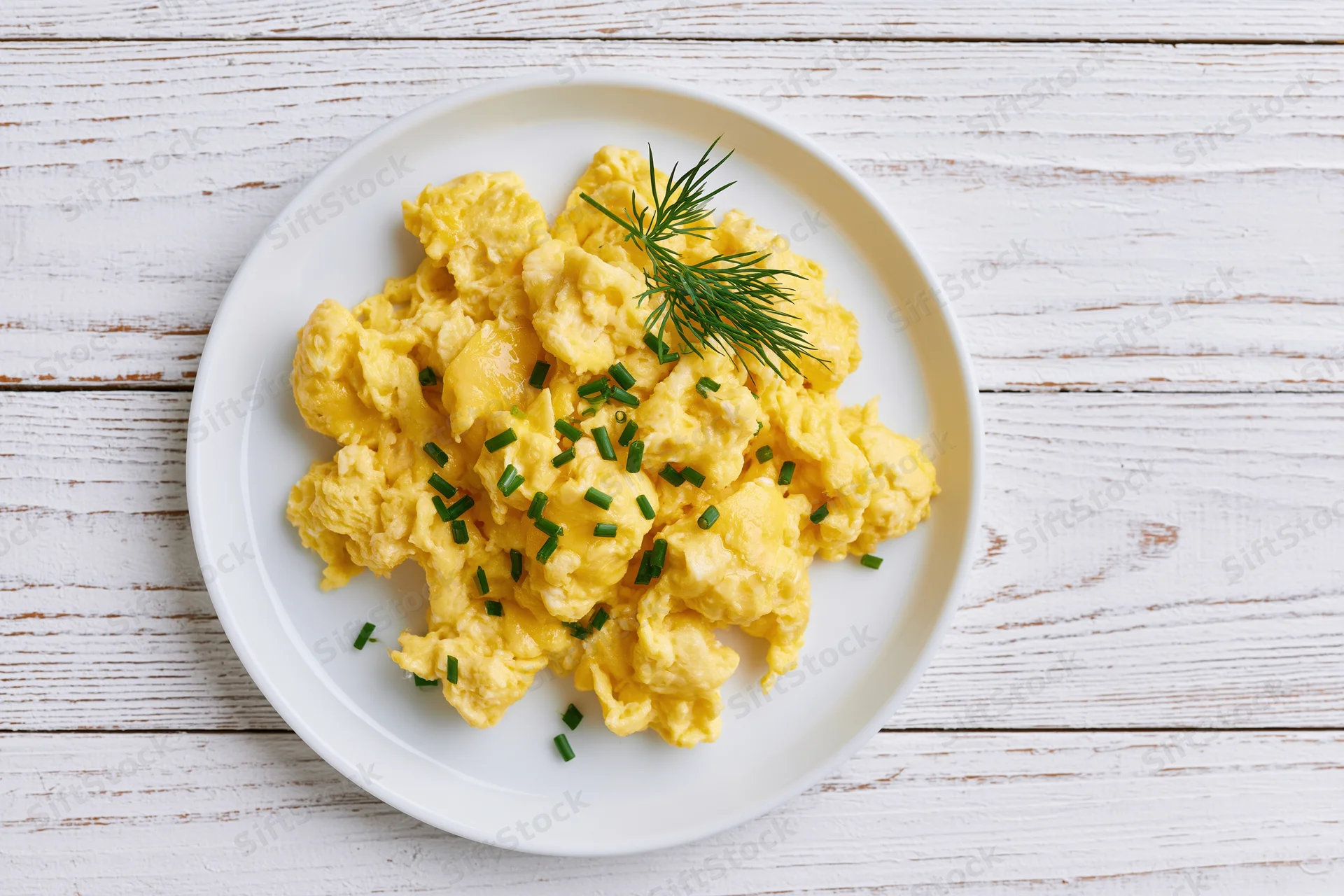 Scrambled eggs garnished with dill and chives on a white ceramic plate set on a wooden table