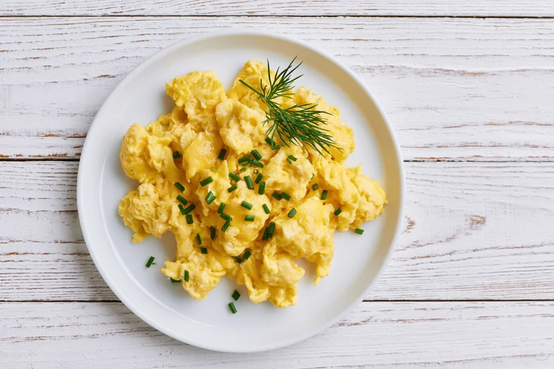 Scrambled eggs garnished with dill and chives on a white ceramic plate set on a wooden table