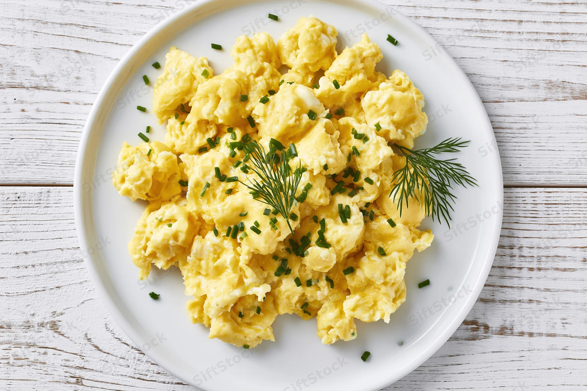 Scrambled eggs garnished with chives and dill on a white ceramic plate set on a wooden surface
