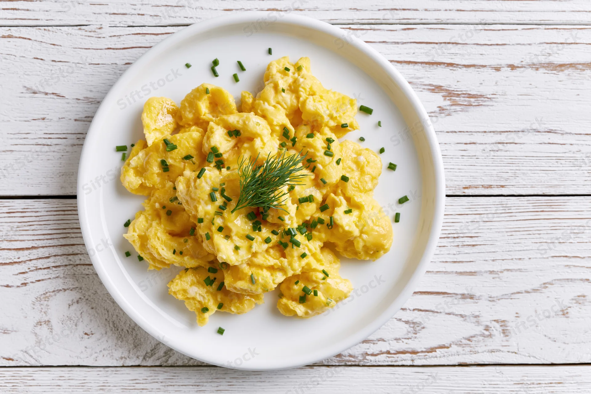 Scrambled eggs piled on a white ceramic plate garnished with chives and dill on a rustic wooden table