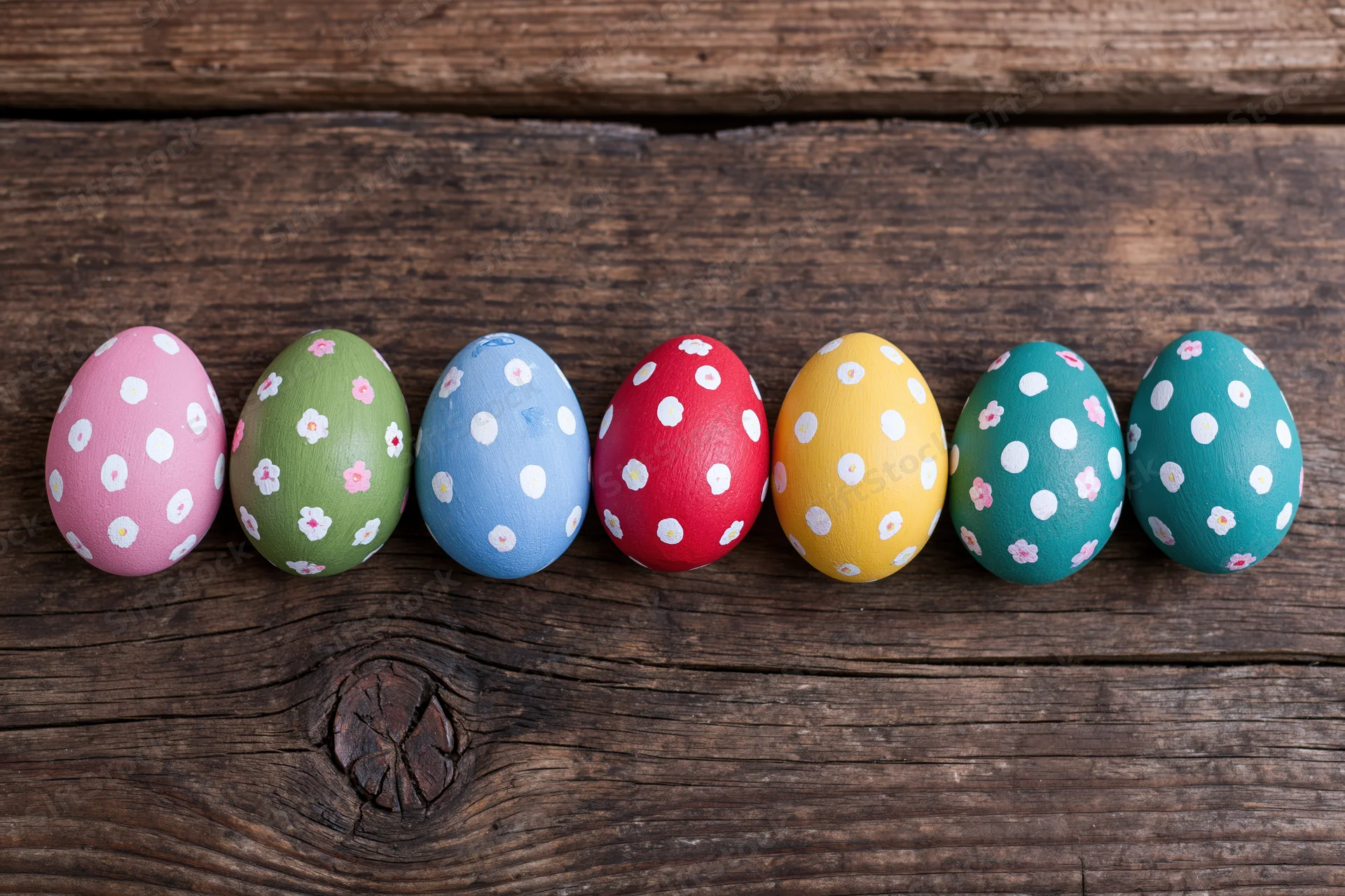Seven hand-painted easter eggs with polka dots and floral patterns arranged in a row on a rustic wooden surface