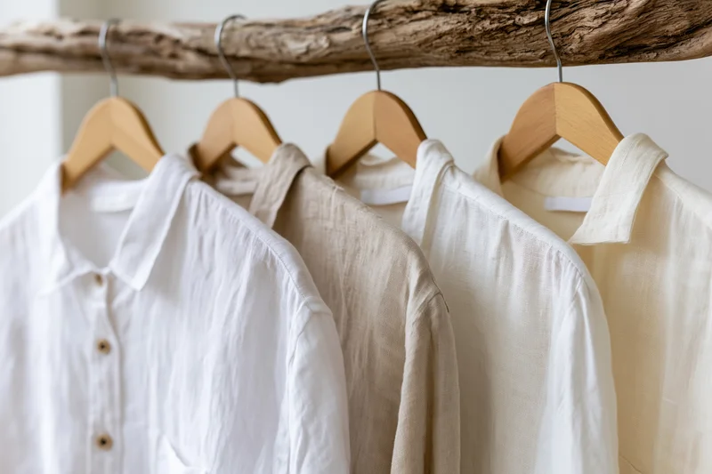 Linen and cotton shirts in white and ecru tones hanging on wooden hangers from a driftwood rack