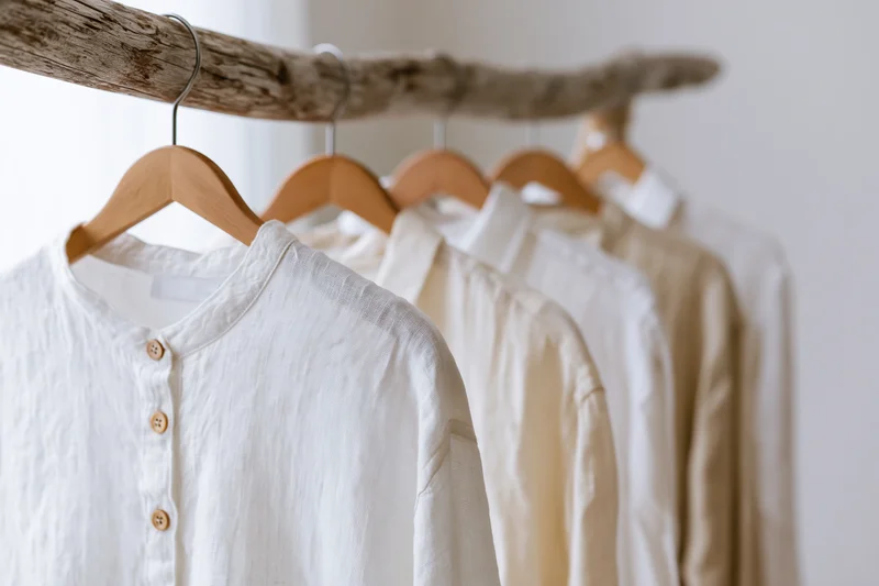 Linen and cotton shirts in white and ecru tones hanging on wooden hangers on a driftwood rack