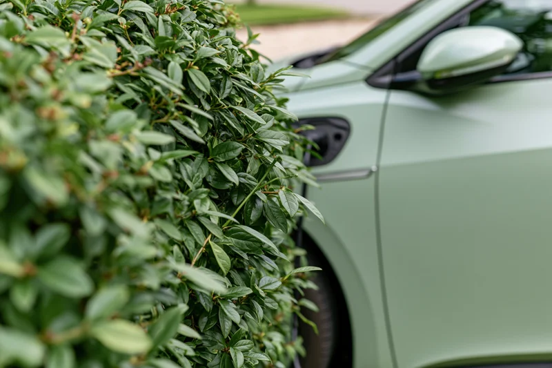 Side profile of a matte sage-green electric vehicle parked beside a leafy green bush in a garden setting