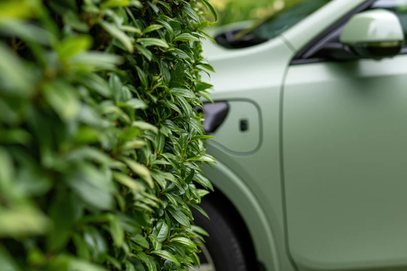 Matte sage-green electric vehicle parked beside a leafy hedge