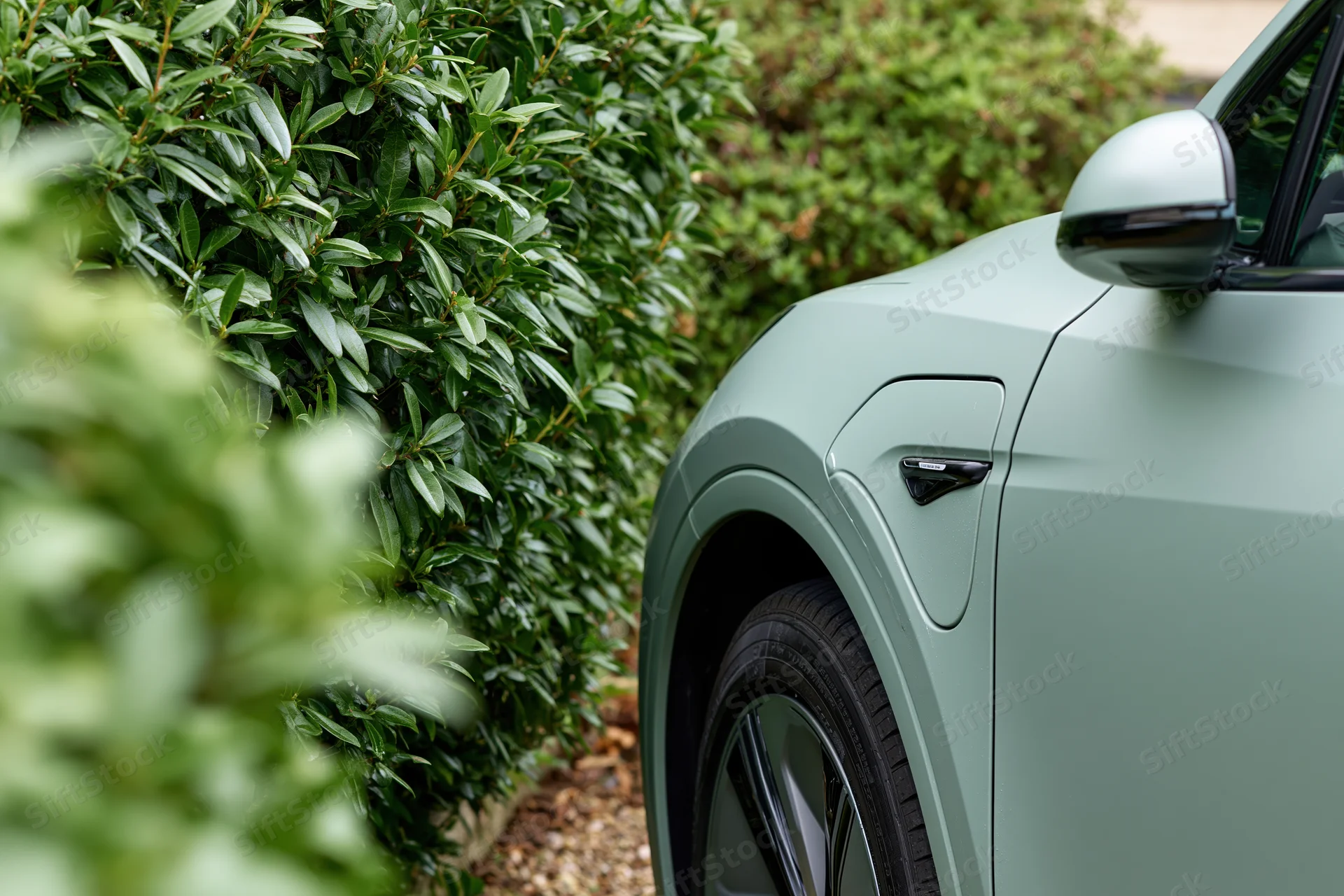 Side profile of a matte sage-green electric vehicle parked beside a leafy hedge in an outdoor garden setting