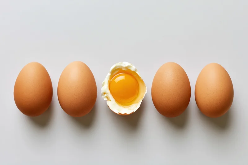 Five whole brown chicken eggs in a row with one cracked open to reveal yolk and white on a flat gray surface
