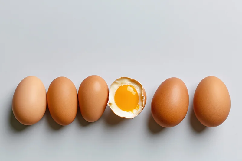 Six brown chicken eggs in a horizontal row on a flat surface with one cracked open revealing yolk and white