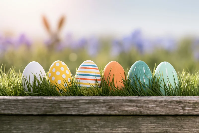 Six painted easter eggs arranged in a row nestled in green grass inside a wooden planter with blurred flowers in the background