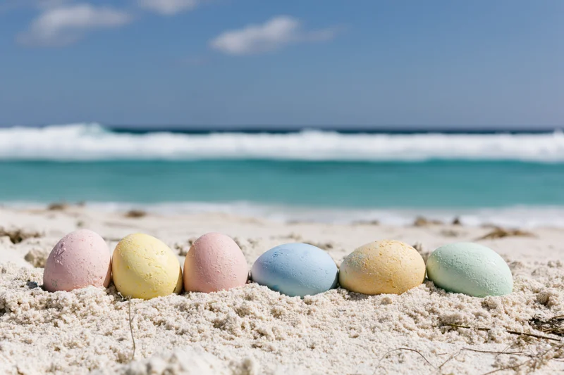 Six pastel-colored easter eggs arranged in a row on white sand at the beach with ocean waves in the background