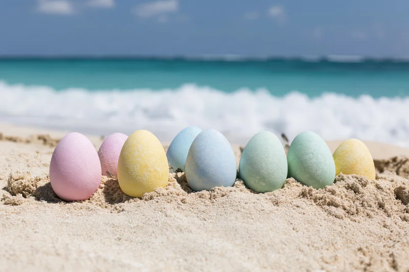 Pastel-colored easter eggs arranged in a row on white sand at the beach with ocean waves in the background