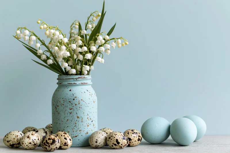 Lily-of-the-valley flowers in a speckled blue mason jar with quail and dyed eggs on a light surface