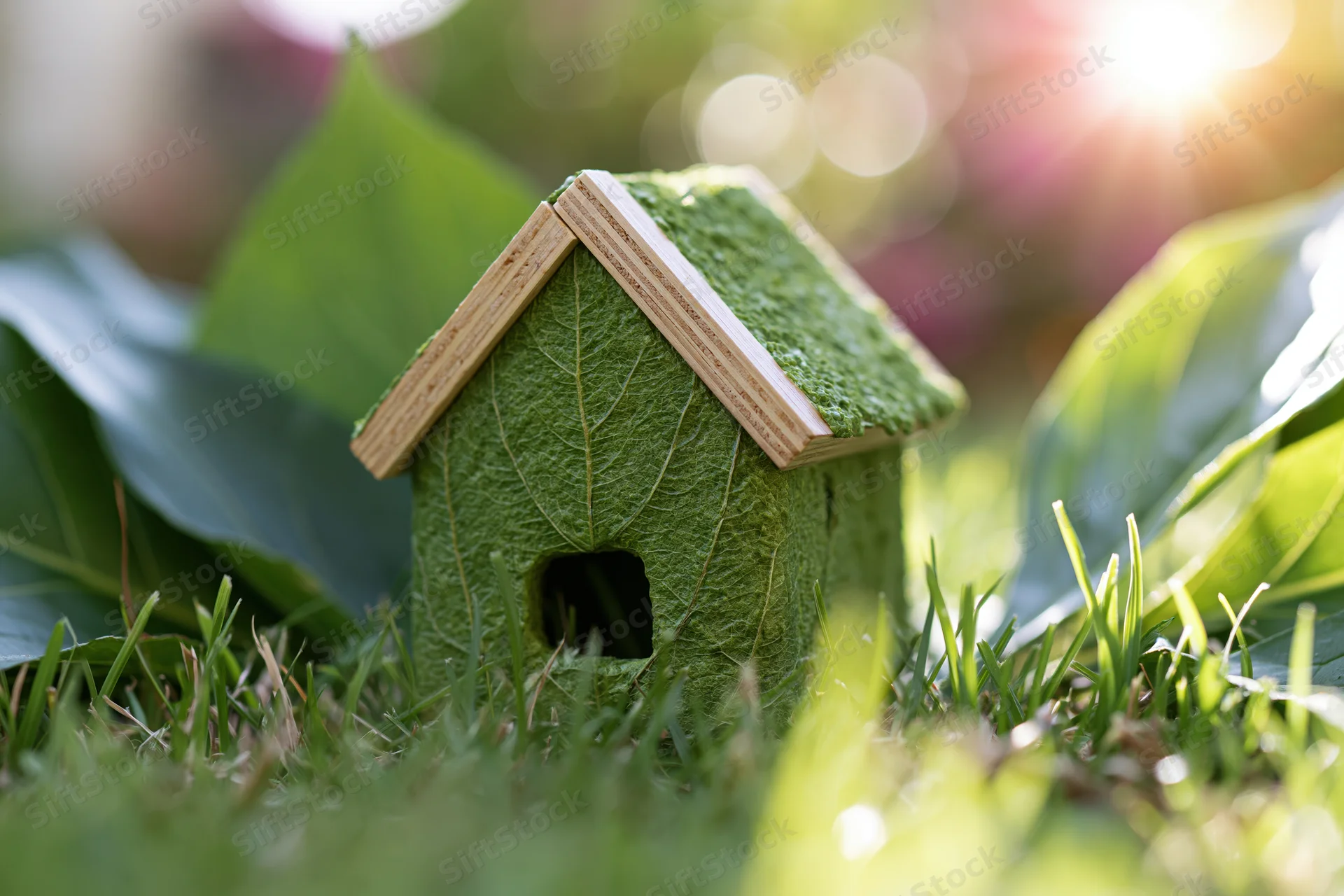 Small green textured house model sitting in fresh grass with blurred foliage and sunlight in background