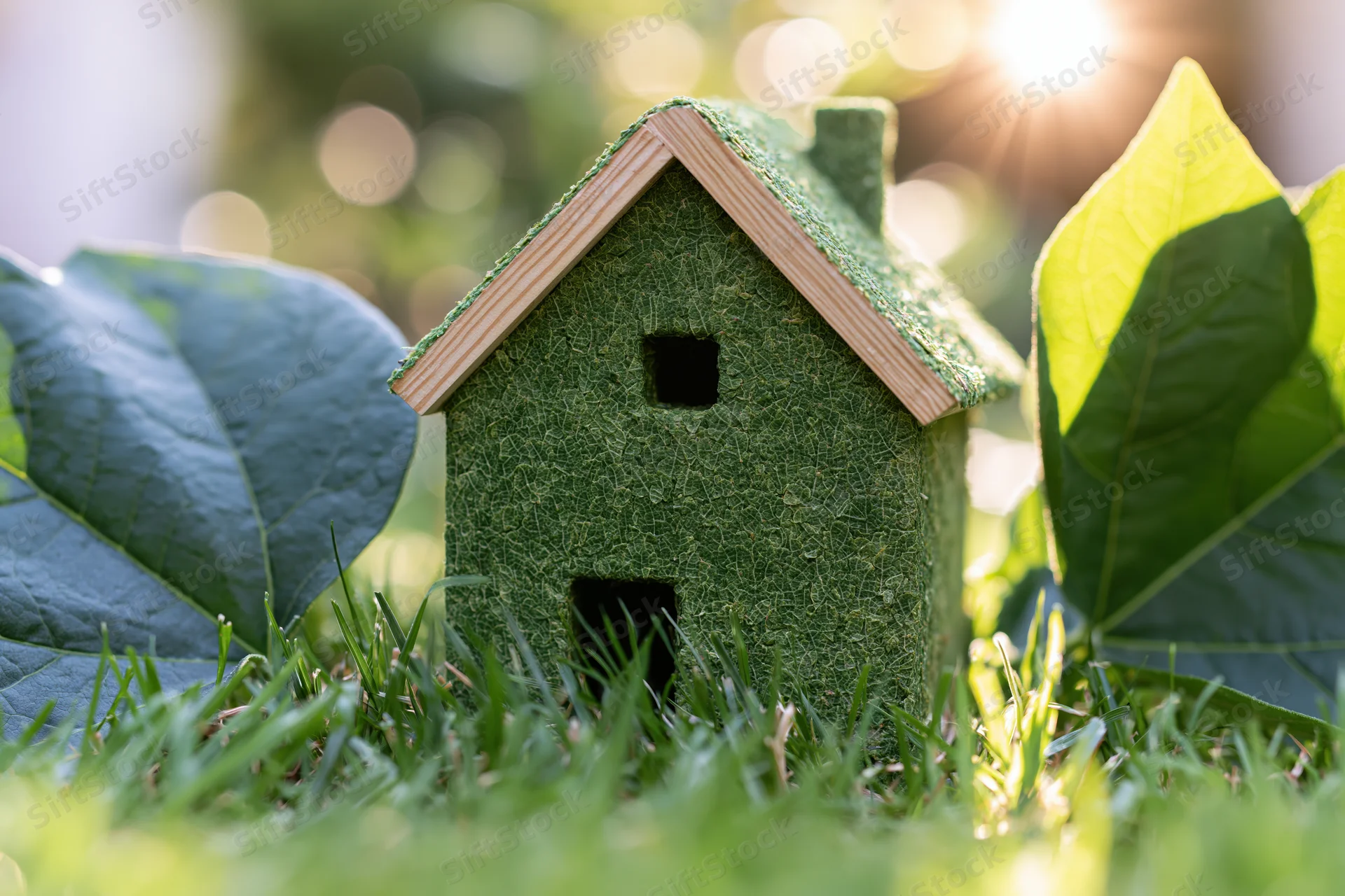 Small green textured house model sitting in fresh grass surrounded by large leaves under sunlight